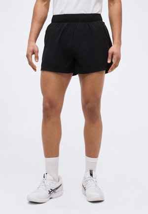 Férfi fekete sportnadrágban, fehér zokniban és fehér futócipőben, egyszerű háttér előtt áll.