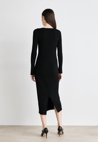 Black ribbed dress with long sleeves, knee-length, featuring a back slit. Worn with black heels, showcasing a sleek and fitted design.