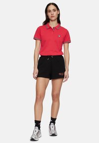 Röd polo tröja med tvåknapps-stängning och randig krage, tillsammans med svarta shorts med dragsko som har en logotypdetalj.