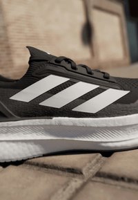Black athletic shoe with white three-stripe design, textured upper, laces, and a white sole with ridged detailing.