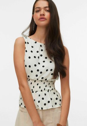 Mujer con cabello largo y oscuro, vistiendo una blusa blanca sin mangas con lunares negros y pantalones beige sobre un fondo liso.