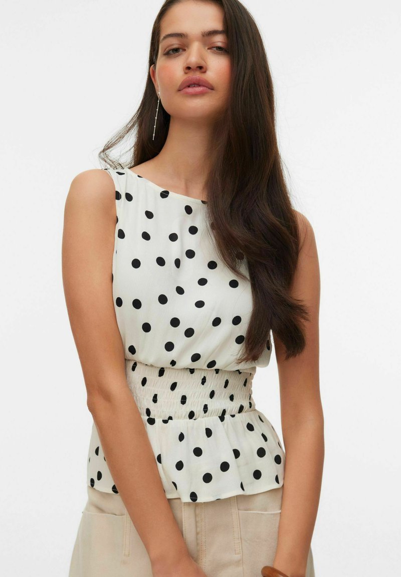 Femme aux longs cheveux foncés portant un chemisier blanc sans manches à pois noirs et un pantalon beige sur un fond uni.