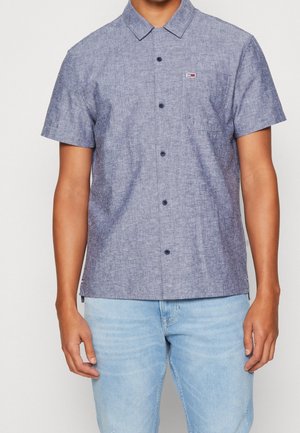Mann trägt ein kurzärmeliges, blaues Chambray-Hemd mit Knöpfen und hellblaue Jeans und steht vor einem einfarbigen Hintergrund.