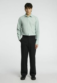 Šviesiai žaliai dryžuota marškinėlių su apykakle, derinami su pritaikytais juodais kelnėmis ir juodais batais, stovintis prieš neutralią fono.