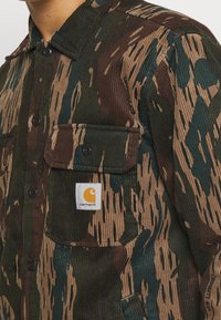 Camicia in corduroy con motivo mimetico che presenta tonalità di marrone, verde e beige. Dettagli notevoli includono due tasche frontali e una toppa con logo.