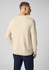 Pull en tricot beige texturé avec un col rond côtelé et des détails à rayures horizontales, associé à un jean bleu foncé. Vue de dos montrée.