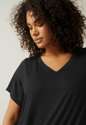 Giovane donna con capelli ricci che indossa una maglietta nera a coste con scollo a V, che guarda verso il basso su uno sfondo chiaro e uniforme.