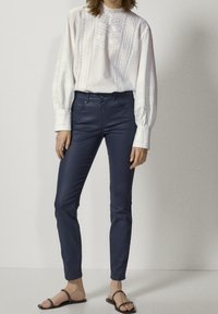 Vit bomullskjorta med intrikat vertikalt broderi, kombinerad med marinblå figurnära byxor. Modellen bär sandaler och visar en avslappnad outfit.