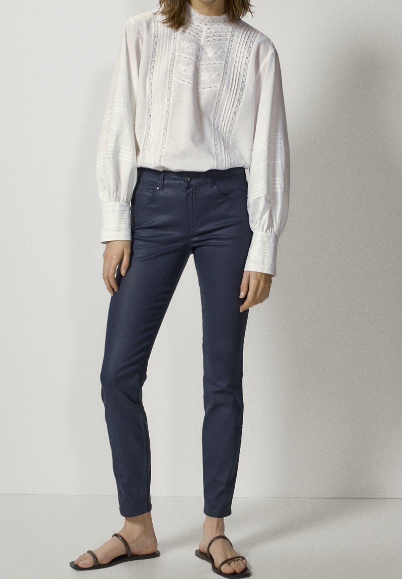 Vit bomullskjorta med intrikat vertikalt broderi, kombinerad med marinblå figurnära byxor. Modellen bär sandaler och visar en avslappnad outfit.