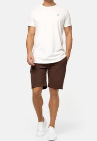 Homme portant un t-shirt blanc uni, un short marron foncé et des baskets blanches, se tenant devant un fond blanc.