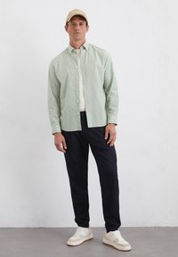 Hellgrün gestreiftes Hemd mit Knöpfen, weißes T-Shirt darunter, marineblaue Hose, beiger Hut und weiße Sneakers mit hellbeigen Akzenten.