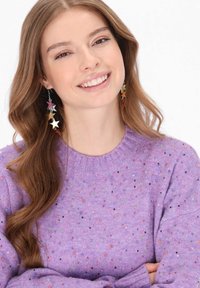 Fioletowy sweter z okrągłym dekoltem, zdobiony kolorowymi plamkami i o miękkiej fakturze. Długie włosy, kolczyki w kształcie gwiazdek w różnych kolorach.