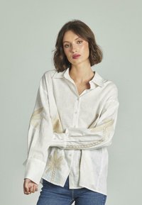 Camicia in lino bianca con ricami floreali dorati sulle maniche e sul davanti. Collo classico, chiusura con bottoni e vestibilità rilassata.