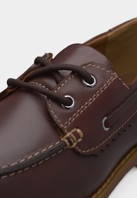 Καφέ δερμάτινο παπούτσι τύπου μοκασίνι με σκούρες καφέ κορδέλες περασμένες από μεταλλικά κρίκους και λεπτομερείς ραφές στα πάνω πλαϊνά και την περιοχή των δαχτύλων.