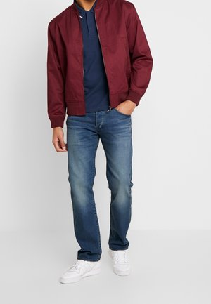 Veste bomber bordeaux, chemise polo marine, jean bleu et baskets blanches. La veste a une fermeture éclair devant et des poignets côtelés.