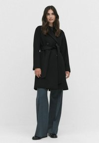 Zwarte trenchcoat met dubbele knopen, een ceintuur in de taille en lange mouwen, gecombineerd met wijde grijze broeken en zwarte schoenen.