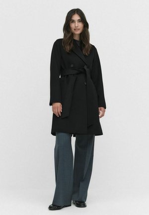 Svart trenchcoat med dubbelknäppta knappar, bälte i midjan och långa ärmar, i kombination med vida gråbyxor och svart fotbeklädnad.