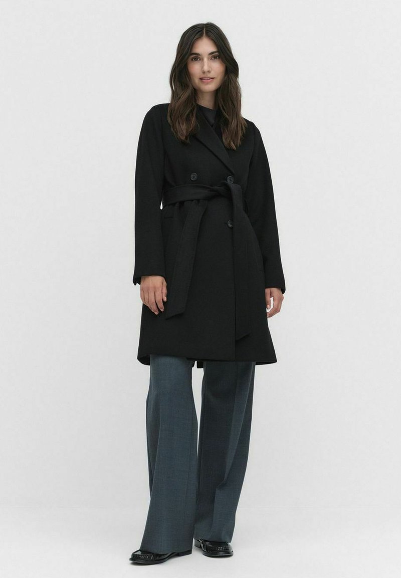 Zwarte trenchcoat met dubbele knopen, een ceintuur in de taille en lange mouwen, gecombineerd met wijde grijze broeken en zwarte schoenen.