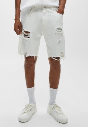 Homme portant un short en jean blanc déchiré jusqu'aux genoux, des chaussettes blanches et des baskets blanches, debout devant un fond uni.