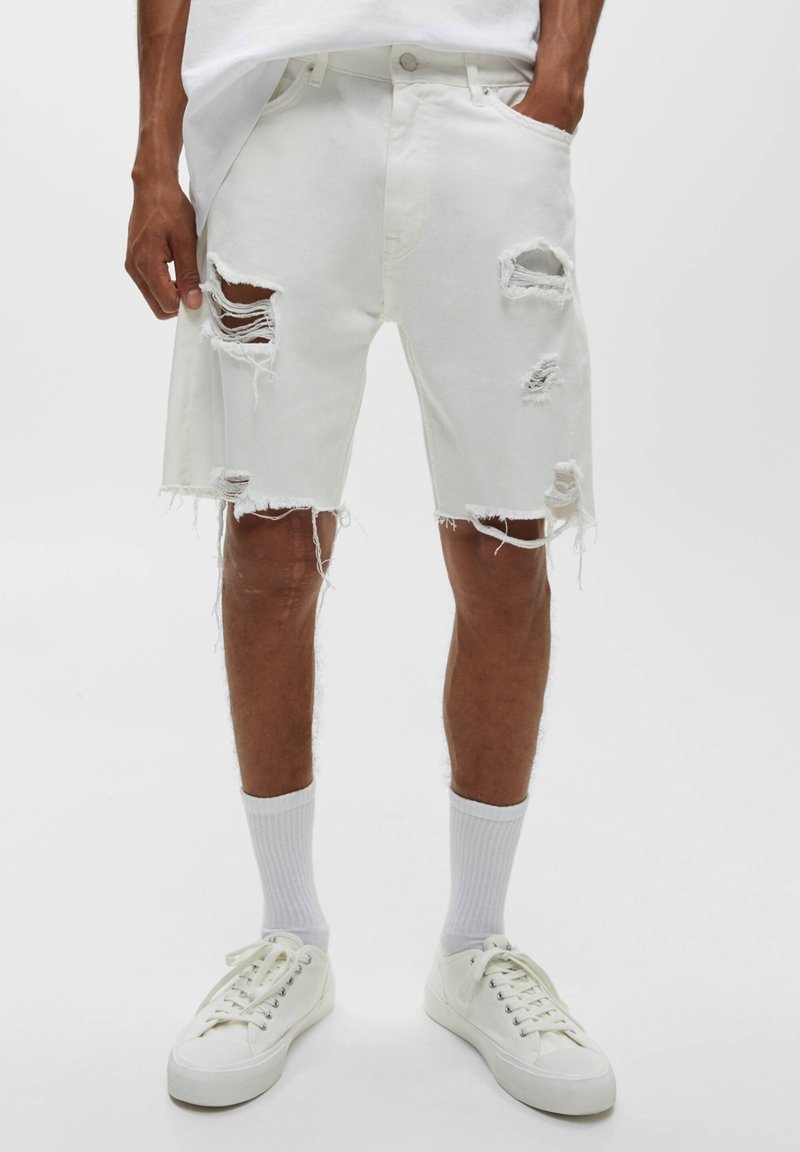 Homme portant un short en jean blanc déchiré jusqu'aux genoux, des chaussettes blanches et des baskets blanches, debout devant un fond uni.