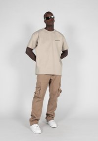Tricou din bumbac bej cu mâneci scurte și logo pe piept, asortat cu pantaloni cargo bej cu buzunare laterale și adidași albi.
