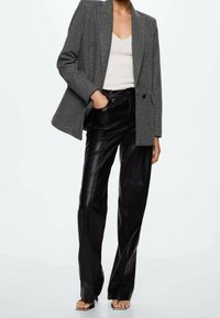 Grijze blazer met patroon en gekartelde revers, gecombineerd met zwarte leren broek met wijde pijpen; het model draagt een witte geribde tanktop en sandalen.