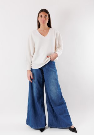 Donna in piedi che indossa un maglione bianco a V, jeans denim blu a gamba larga e scarpe nere appuntite, su uno sfondo bianco semplice.