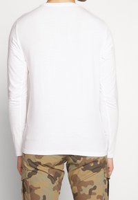 Valkoinen pitkähihaiseen paita, joka on valmistettu pehmeästä puuvillasta. Pyöreä pääntie, slim fit -mallinen, jossa on minimumisuojaus. Yhdistetty camo-housujen kanssa.