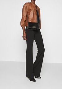 Person som har på sig en brun kort skinnjacka, svarta byxor, svarta spetsiga skor och en svart axelväska med kedjestropp.