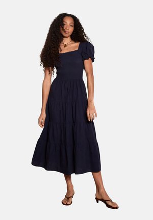 Kobieta z długimi kręconymi włosami, ubrana w granatową sukienkę midi z falbankami i bufkami oraz brązowe sandały na obcasie, stojąca na białym tle.