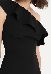 Schwarzes, einseitig tragbares Kleid mit großen Rüschendetails am Halsauschnitt. Der Stoff wirkt glatt und hat eine taillierte Silhouette.