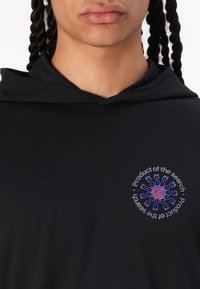 Haut à capuche noir avec un design graphique rond en bleu et rouge sur la poitrine, comportant le texte "Produit de la recherche." Matériel en coton doux.