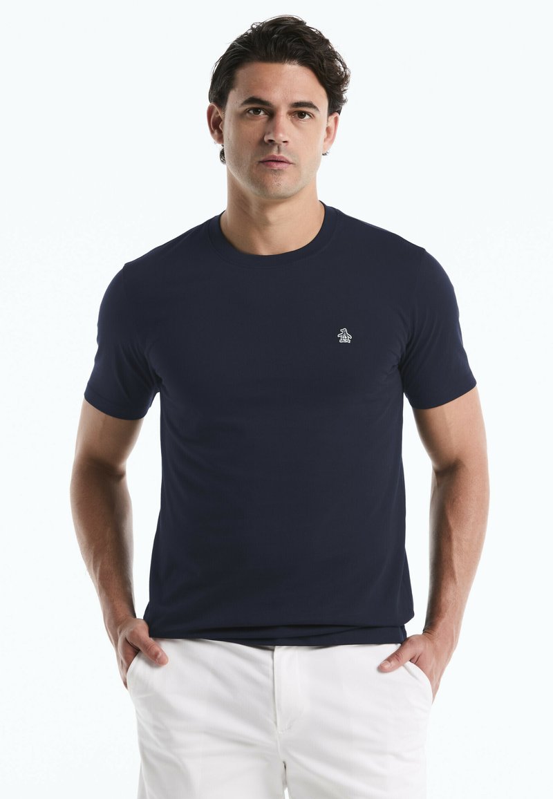 Homme portant un T-shirt bleu marine ajusté avec un logo de manchot sur la poitrine et un pantalon blanc, debout les mains dans les poches sur un fond blanc.