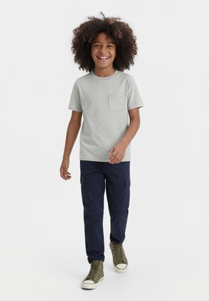 Usmievajúce sa dieťa s kučeravými vlasmi kráča vpred, oblečené v šedej tričku s vreckami, námorníckych cargo nohaviciach a olivovo zelených vysokých teniskách.