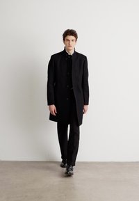 Palton negru din lână cu guler înalt și nasturi, purtat peste pantaloni negri și pantofi. Design minimalist cu o croială ajustată.