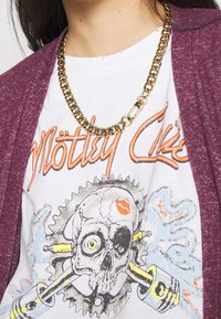 Un collar de cadena de oro se desliza sobre una camiseta gráfica blanca con un cráneo y colores brillantes, combinada con un cárdigan texturado en color marrón burdeos.