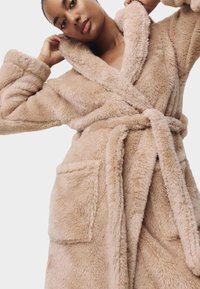 Donna che sistema il colletto di una vestaglia beige, morbida, con cintura e grandi tasche applicate, su uno sfondo neutro.
