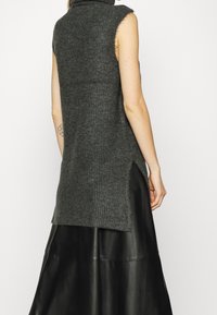 Pull sans manches gris avec un col montant, présentant une texture côtelée et des fentes latérales, associé à une jupe en cuir noire.