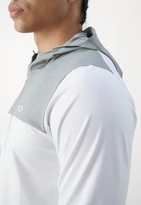 Haut de hoodie sportif pour homme avec un dessus texturé gris et des manches blanches, présentant une fermeture éclair à l'avant et un design ajusté. Matériau léger et respirant.