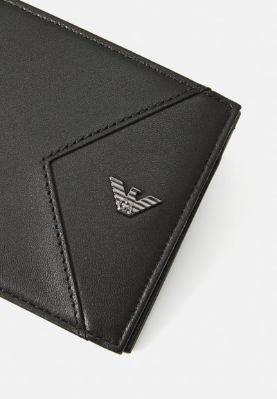 Cartera de cuero negro con una superficie texturizada, diseño angular, acentos cosidos y un emblema de logo plateado en la esquina inferior.