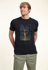 Svart bomulls t-shirt med en färgglad grafisk tryck av en filmaffisch för "Highlander," med fet röd text och detaljerad bakgrundsdesign.