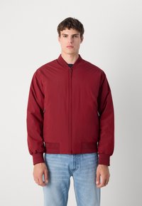 Red bomber jacket with a round collar, zip closure, and ribbed cuffs. Features a smooth, lightweight fabric and side pockets.