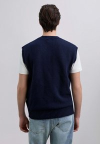 Suéter sin mangas de punto azul marino con dobladillo acanalado, usado sobre una camiseta blanca; modelo de espaldas, mostrando los detalles y la textura de la parte posterior.