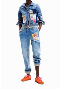 Person som bär en denimjacka och joggbyxor med färgglada Mickey Mouse-lappar, tillsammans med klara orangea sneakers.