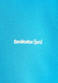 T-shirt turquoise en tissu doux présente le texte "Smilodox Sport" en blanc, centré et légèrement courbé.