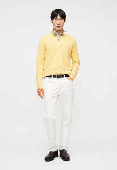 Άνδρας που φοράει κίτρινο πουλόβερ με φερμουάρ στο στήθος πάνω από ριγέ πουκάμισο, λευκό παντελόνι, μαύρη ζώνη και σκούρα καφέ μπότες, στέκεται μπροστά σε απλό φόντο.
