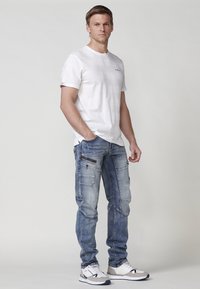 Camicia bianca maniche corte con sottili righe, abbinata a pantaloni cargo in denim azzurro chiaro con molteplici tasche e dettagli in zip scuri.