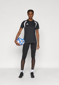 Μαύρο αθλητικό μπλουζάκι με λευκές λεπτομέρειες, σε συνδυασμό με μαύρο κολάν με πλευρικές ρίγες. Το άτομο κρατάει μια πολύχρωμη ποδοσφαιρική μπάλα.