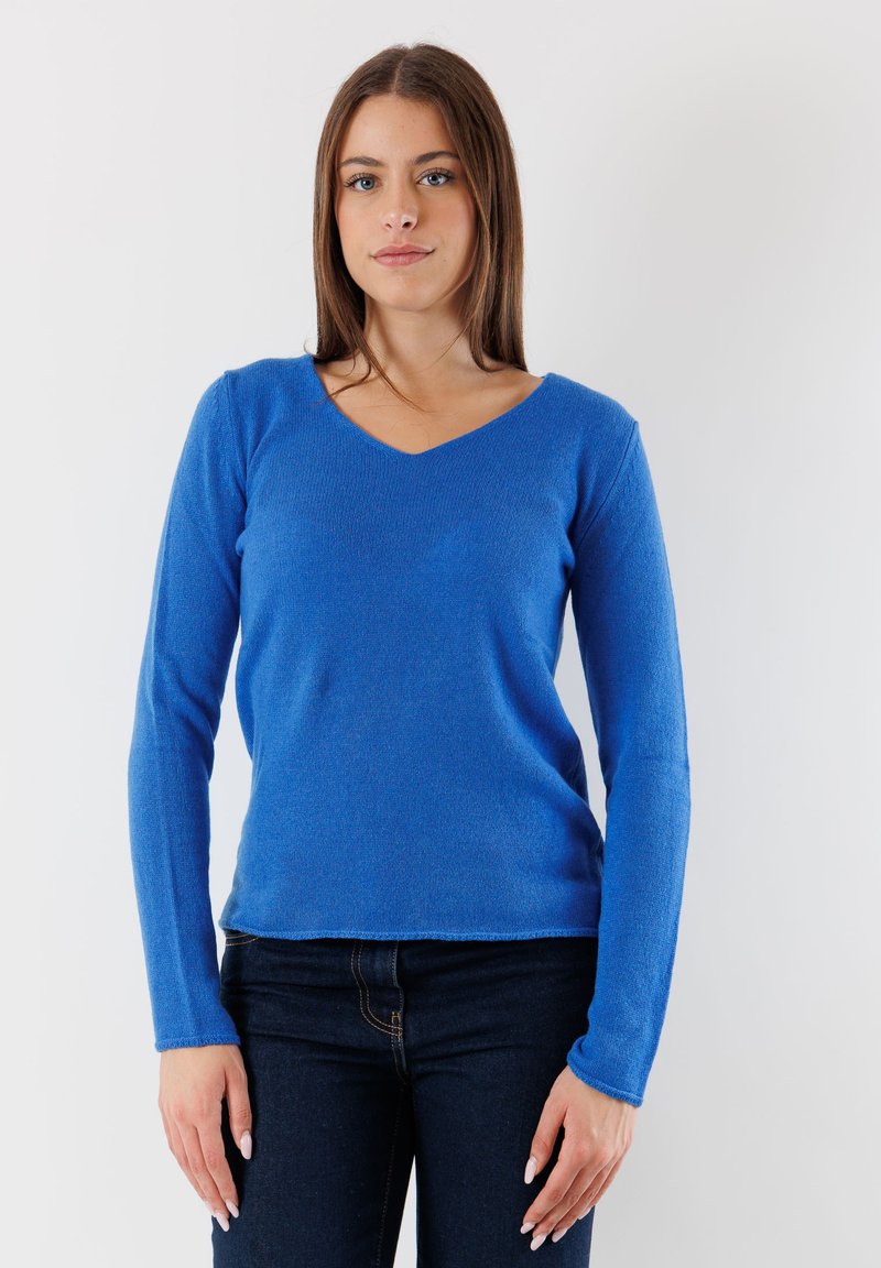 Giovane donna in piedi con un maglione blu a maniche lunghe e scollo a V e jeans scuri, contro uno sfondo bianco semplice.