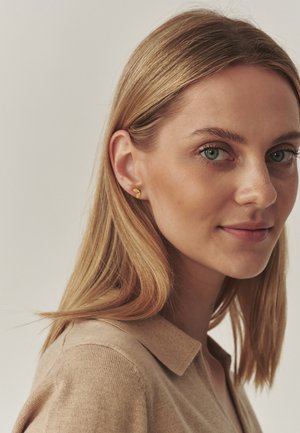 Boucles d'oreilles en or avec un design courbé en forme de feuille. Le modèle a les cheveux châtain clair et porte un pull beige à col, faisant légèrement face à la gauche.
